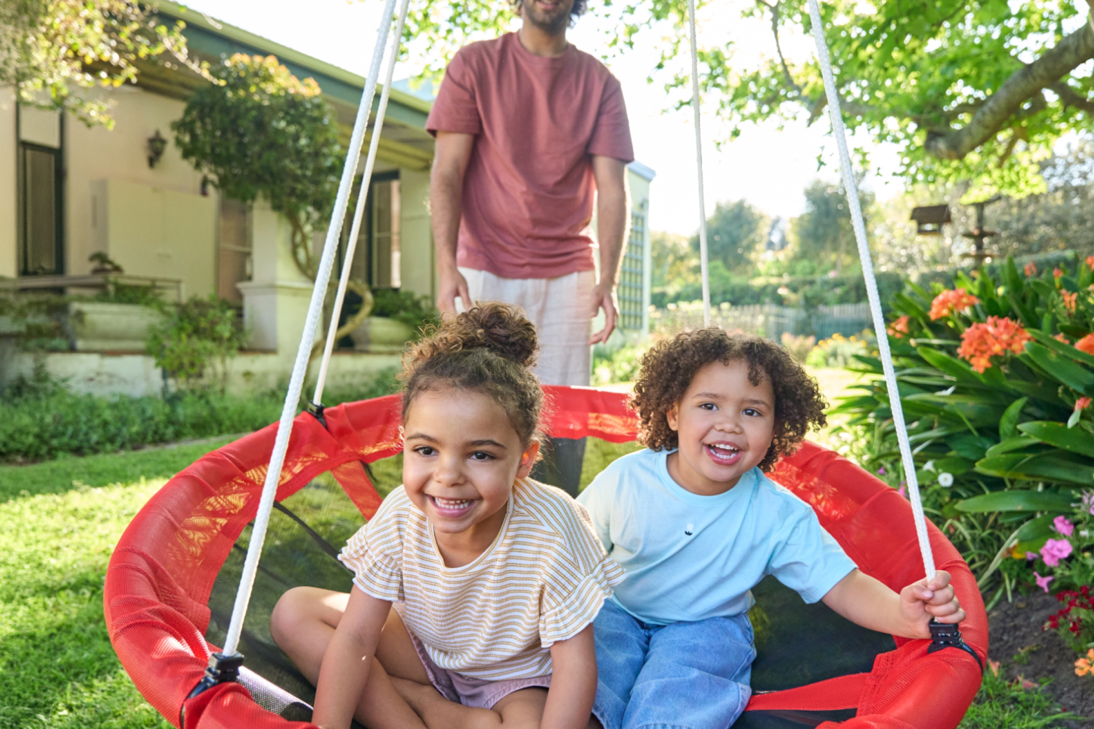 Dwoje dzieci siedzi na czerwonej huśtawce, a dorosły stoi za nimi w ogrodzie.