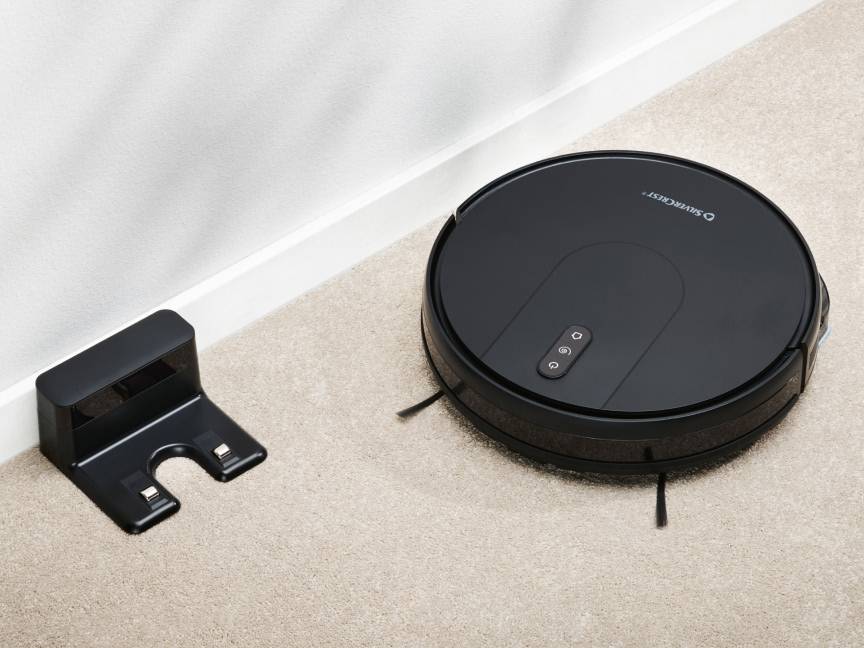 Czarny robot odkurzający i stacja dokująca na beżowym dywanie przy białej ścianie.