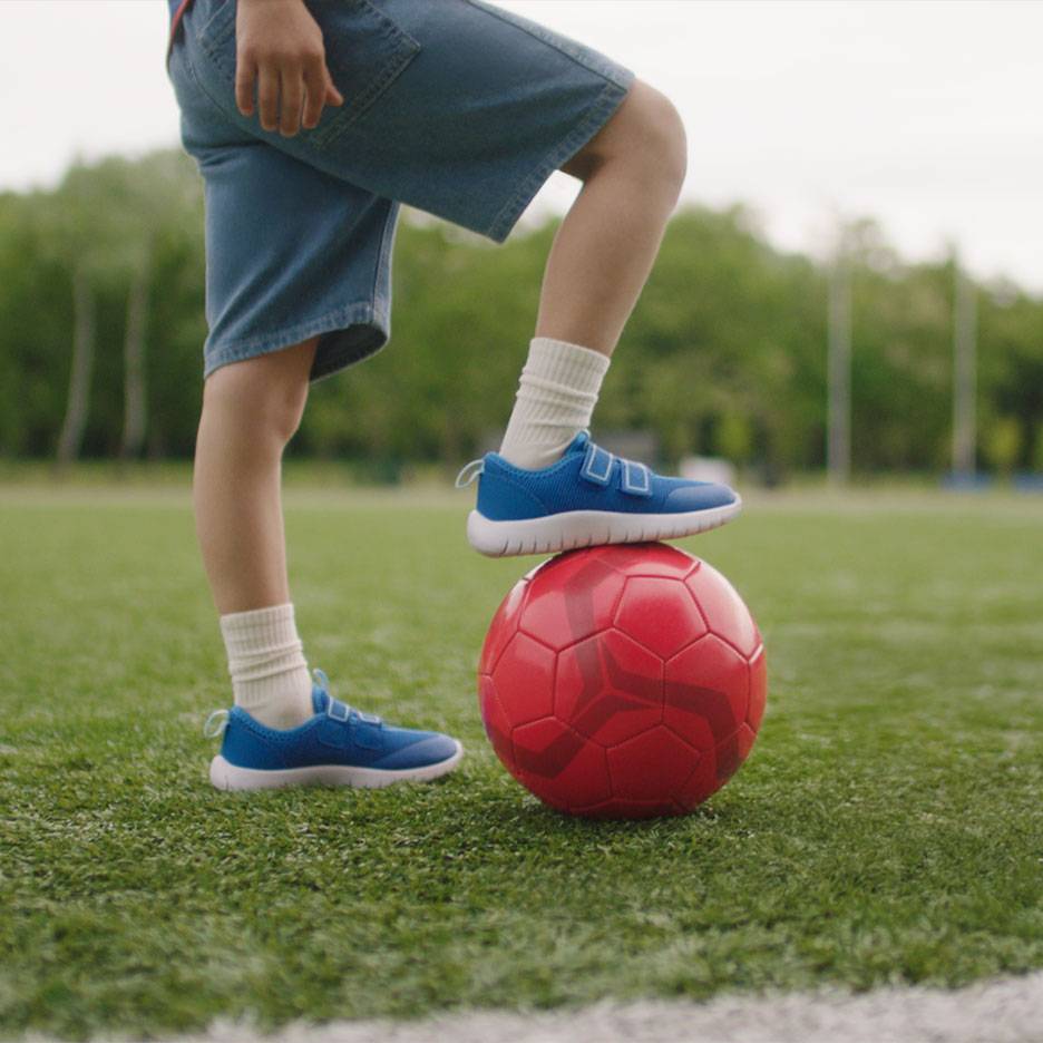 Dziecko w szortach i niebieskich sneakersach, z jedną stopą na czerwonej piłce nożnej.