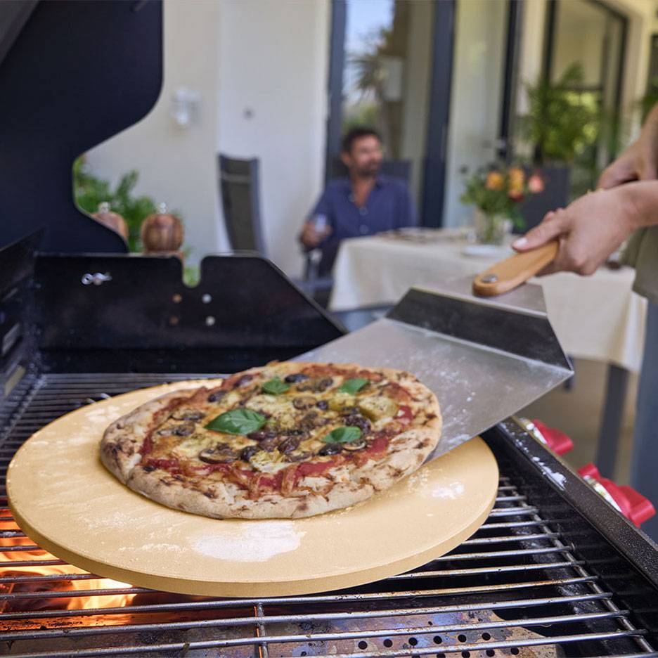 Pizza na kamieniu do pizzy na grillu, z łopatką i mężczyzną w tle.