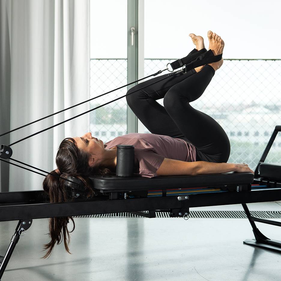 Kobieta ćwiczy nogi na reformerze Pilates z taśmami oporowymi.
