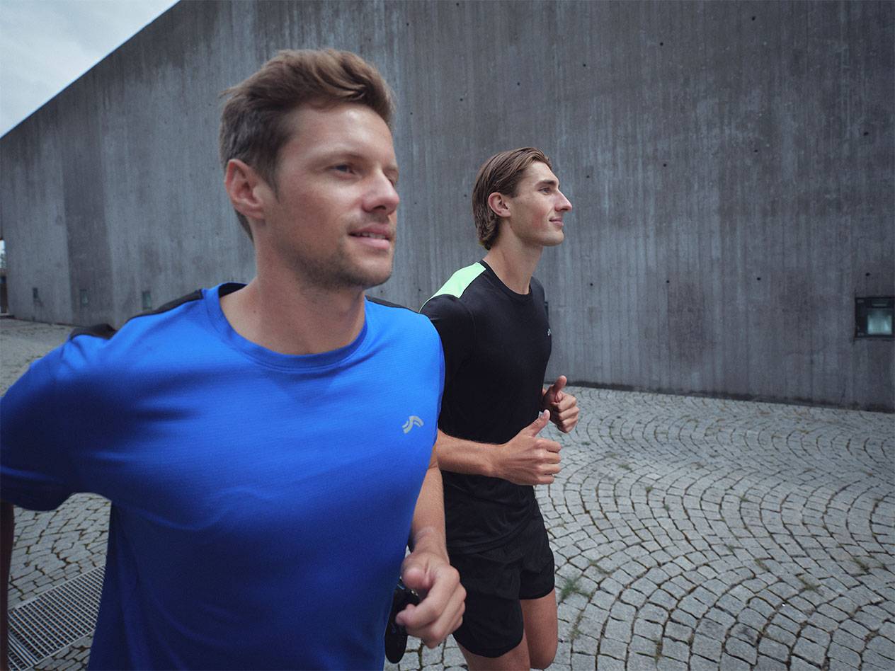Dwóch mężczyzn biegnie w sportowych koszulkach i szortach, jeden w niebieskiej, drugi w czarnej.