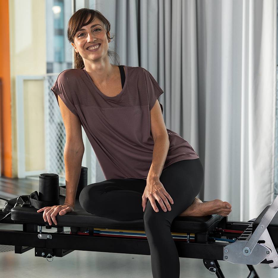 Kobieta w stroju sportowym uśmiecha się, siedząc na maszynie do pilatesu.