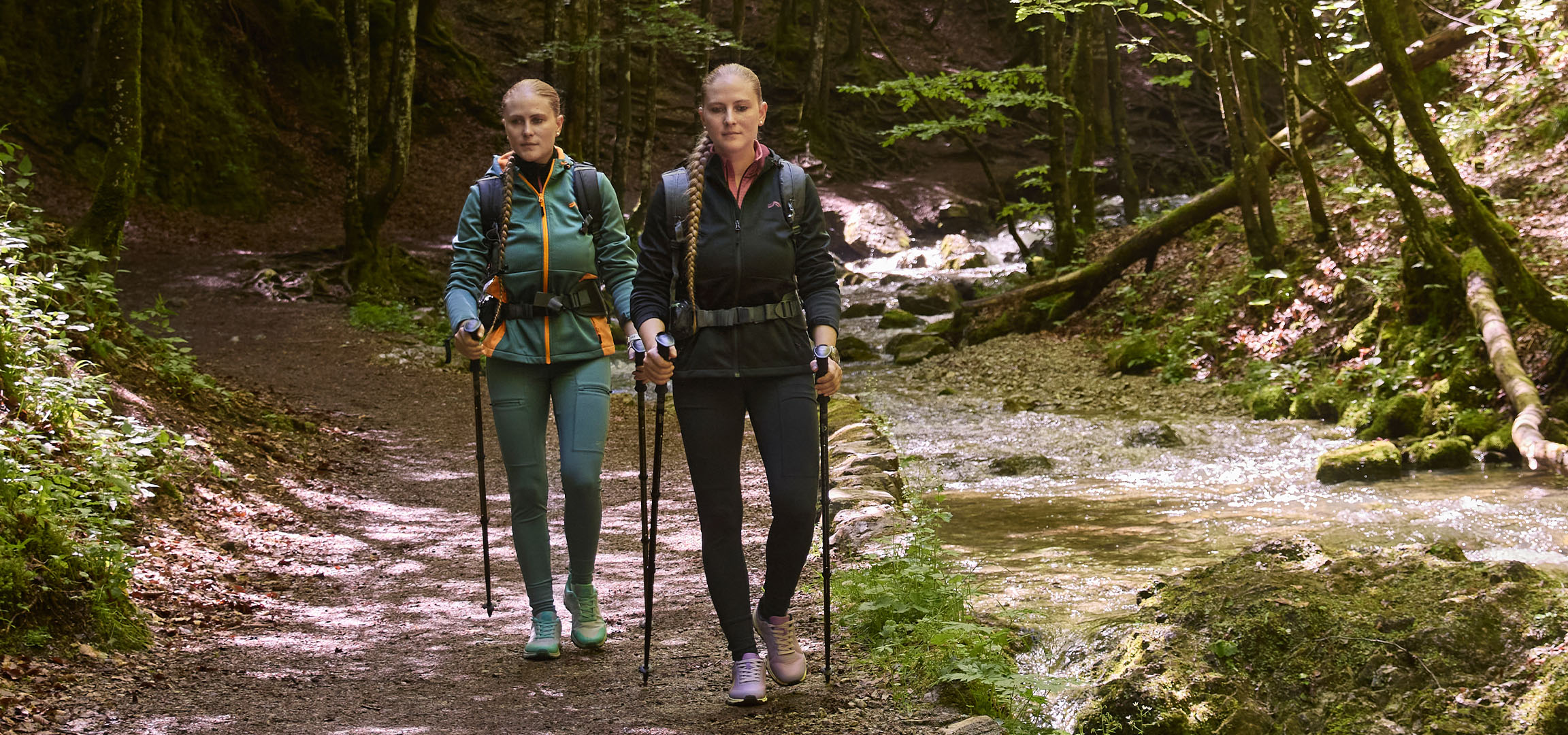 Dwie kobiety wędrują wzdłuż strumienia w lesie z kijkami trekkingowymi.