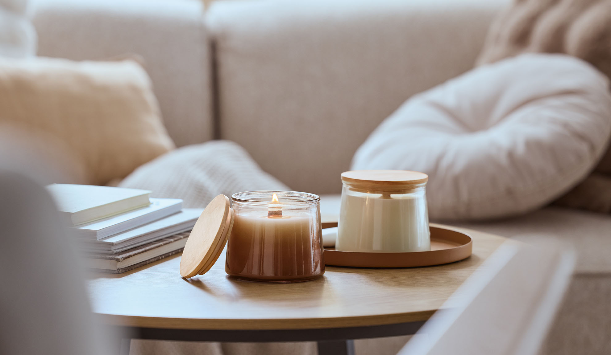 Dwie świece zapachowe na drewnianym stole, jedna zapalona, z przytulną sofą w tle.