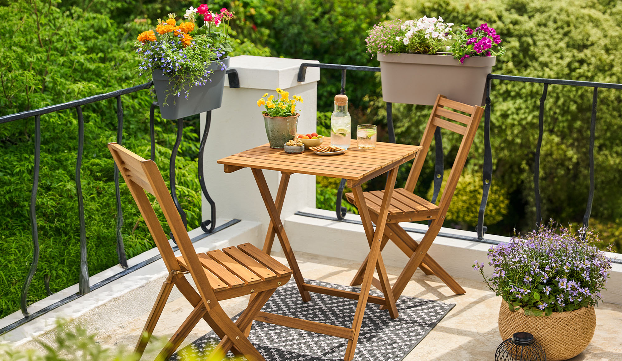Balkon z drewnianym zestawem bistro, kwiatami w doniczkach i napojami na wzorzystym dywanie.
