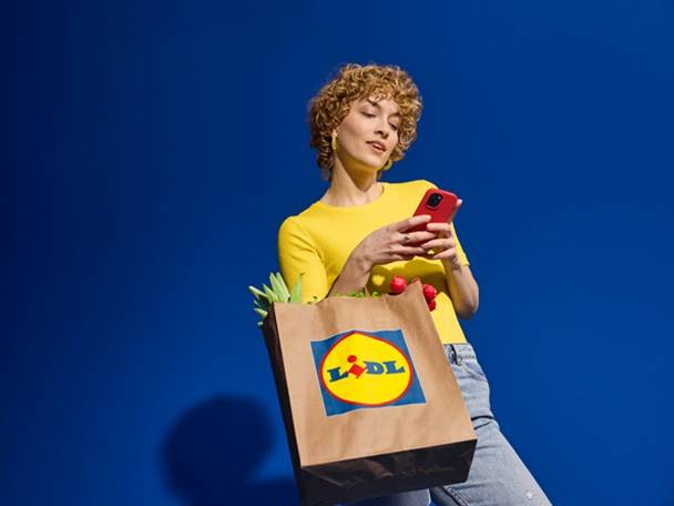 Kobieta w żółtej koszulce trzymająca telefon i torbę na zakupy z logo Lidl.