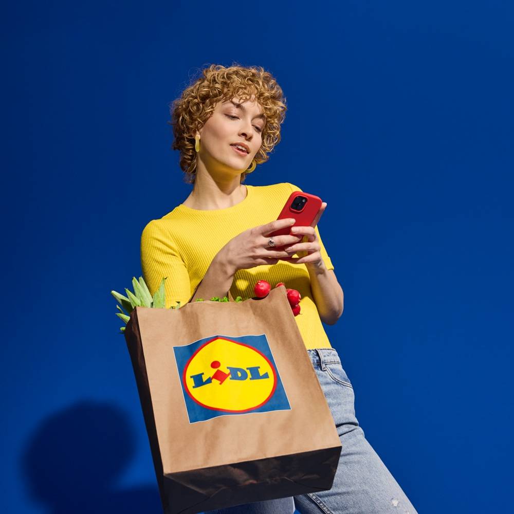 Kobieta w żółtej koszulce trzyma torbę na zakupy Lidl z warzywami i czerwony smartfon.