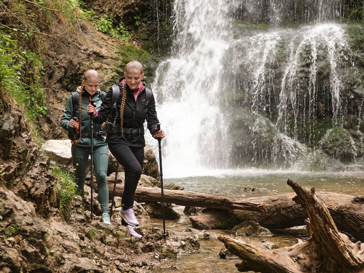Dwie kobiety z kijkami trekkingowymi idą skalistą ścieżką w pobliżu wodospadu.